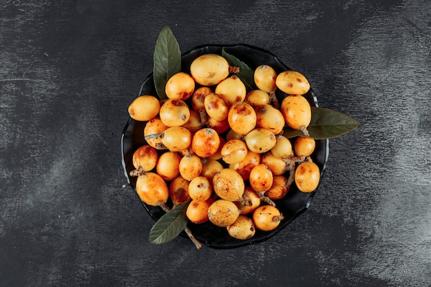 Loquat con le foglie in una ciotola su fondo strutturato scuro, vista superiore.