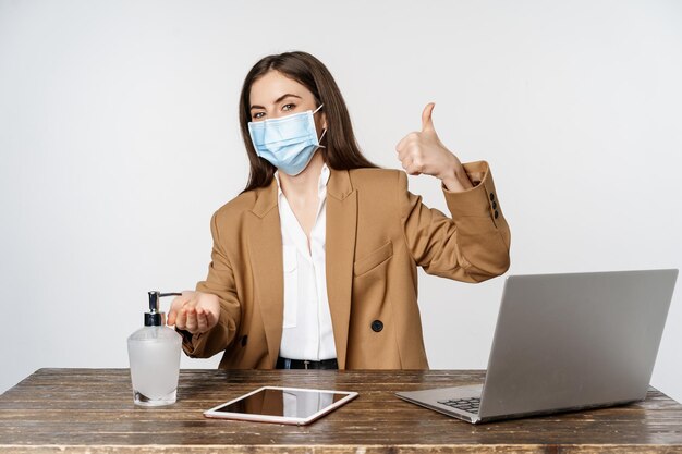 Luogo di lavoro e concetto covid-19. Donna d'affari sorridente con maschera medica facciale, che mostra i pollici in su mentre si utilizza un detergente disinfettante per le mani per pulire le mani lavate, sfondo bianco.