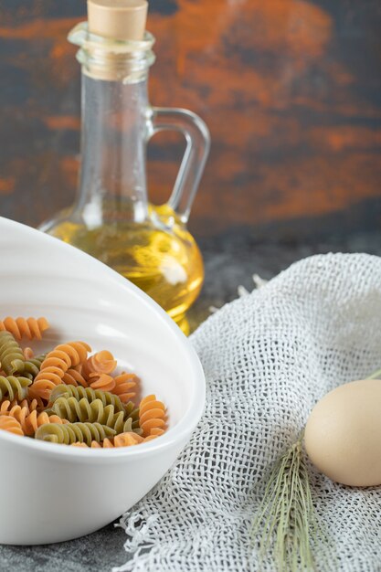 Maccheroni a spirale crudi nel piatto bianco con l'uovo e la bottiglia di olio