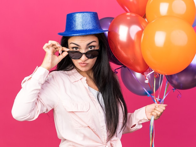 Macchina fotografica dall'aspetto fiducioso giovane bella ragazza che indossa cappello da festa e occhiali che tengono palloncini