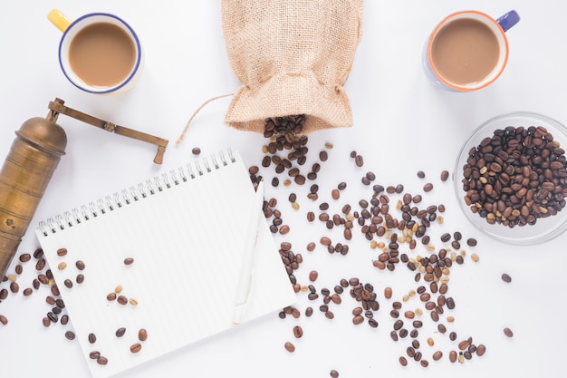 Macinino da caffè vecchio; chicchi di caffè; tazza di caffè; Blocchetto per appunti a spirale in bianco con la penna su priorità bassa bianca