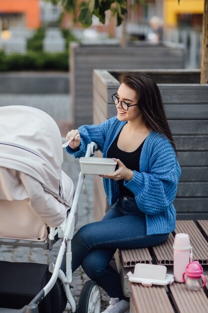 Madre che cammina con il neonato nel passeggino donna che tiene in mano una scatola di plastica e dà da mangiare al suo bambino