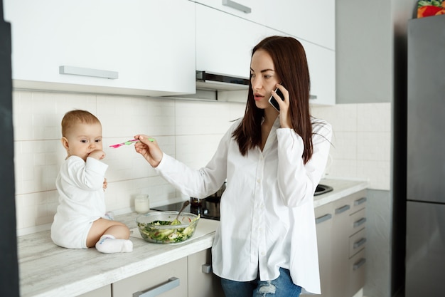 Madre che lavora da casa, parla al telefono e nutre un bambino piccolo e carino