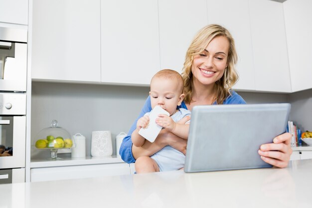 Madre che tiene bambino mentre si utilizza tavoletta digitale in cucina