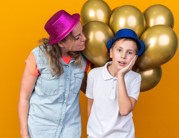 madre contenta che indossa un cappello da festa viola e tiene in mano palloncini di elio guardando suo figlio sorpreso con un cappello da festa blu che mette la mano sul viso isolato sulla parete arancione con spazio di copia