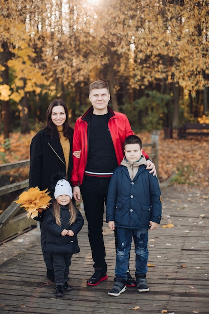 Madre di famiglia felice, padre e due bambini che posano insieme all'aperto al parco autunnale a pieno regime. Bambino sorridente e genitori che camminano insieme tenendo in mano foglie gialle che provano emozioni positive