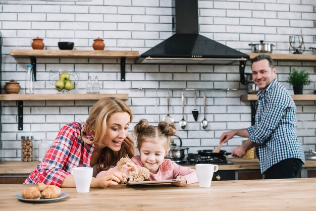 Madre di insegnare a sua figlia di utilizzare la tavoletta digitale mentre padre preparare il cibo in cucina