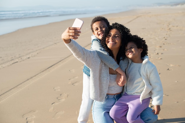 Madre e bambini che prendono selfie sulla spiaggia. Famiglia afroamericana che trascorre del tempo insieme all'aria aperta, scattando foto con il telefono cellulare. Tempo libero, social media, concetto di genitorialità