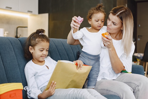 Madre e bambini che si rilassano insieme sul divano di casa in soggiorno. Bambine che leggono un libro e giocano a giocattoli.