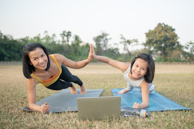 Madre e figlia che fanno yoga. formazione donna e bambino nel parco. sport all'aperto. stile di vita sportivo sano, posa chaturanga. benessere, concetto di consapevolezza, guardare video tutorial online sul laptop