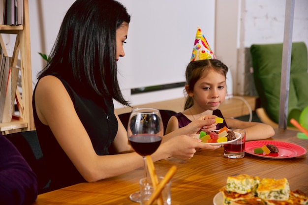 Madre e figlia che festeggiano un compleanno a casa. Grande famiglia che mangia torta e beve vino mentre saluta e si diverte i bambini. Celebrazione, famiglia, festa, casa, infanzia, concetto di genitorialità.