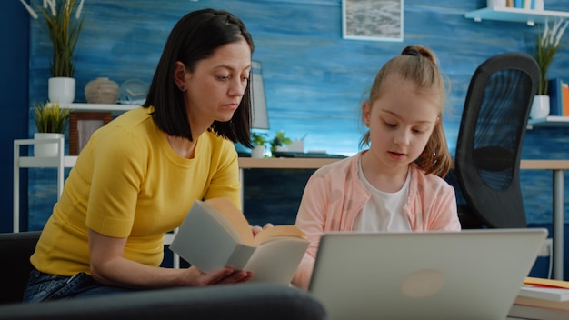 Madre e figlia che imparano insieme a libri e laptop per l'istruzione a distanza. Bambino che utilizza il dispositivo per studiare per corsi e lezioni online. Ragazza della scuola elementare che studia con il genitore