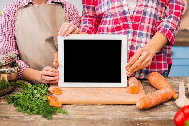 Madre e figlia che mostrano compressa digitale dello schermo in bianco sul tagliere con le verdure