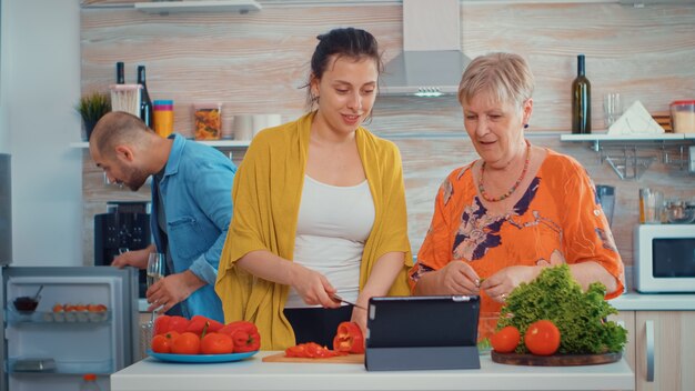 Madre e figlia che tagliano il pepe e guardano nel tablet, cucinando utilizzando la ricetta del tablet digitale online sul computer del pc nella cucina di casa. durante la preparazione del pasto. Fine settimana rilassante e accogliente per famiglie allargate