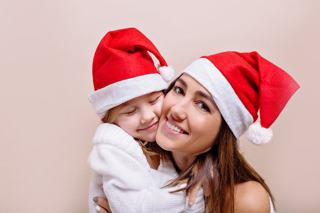 Madre e figlia divertenti felici e sorridenti stanno posando e tenendo i loro volti indossando i cappelli di Babbo Natale. Una giovane bella donna con labbra luminose tiene in mano una bambina di 5 anni.