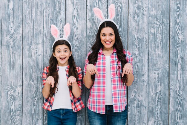 Madre e figlia emozionanti con le orecchie del coniglietto che indicano il dito verso il basso contro il contesto di legno