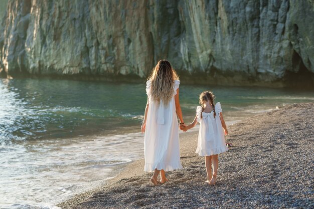 Madre e figlia felici in vestito bianco che camminano nella spiaggia durante il tramonto. vista posteriore