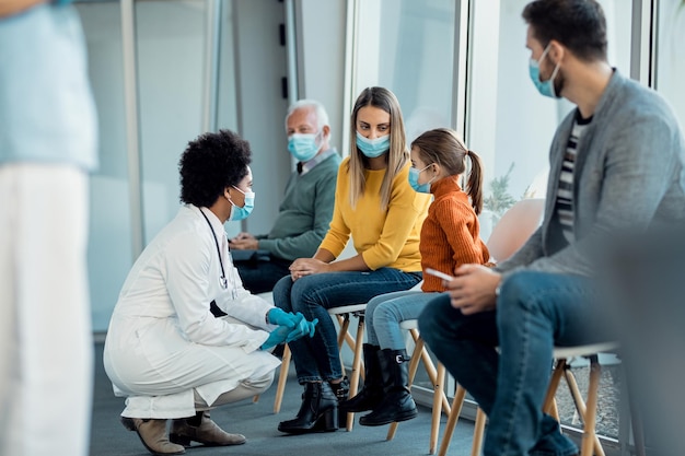 Madre e figlia parlano con un medico afroamericano mentre indossano maschere facciali nella sala d'attesa dell'ospedale