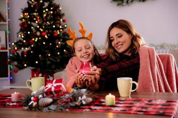 Madre e figlia sorridenti che tengono e che esaminano candela che si siedono al tavolo godendo il tempo di natale a casa