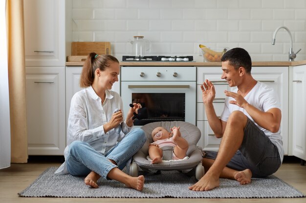 Madre e padre che giocano con il figlio o la figlia sulla sedia a dondolo sul pavimento della stanza leggera con la cucina sullo sfondo, famiglia felice che trascorre del tempo insieme, giocando con il bambino.