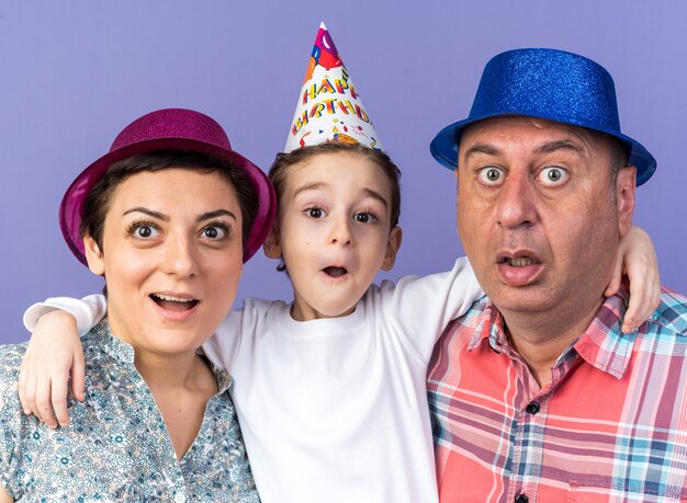 madre e padre sorpresi con cappelli da festa in piedi con il figlio isolato sul muro viola con spazio copia