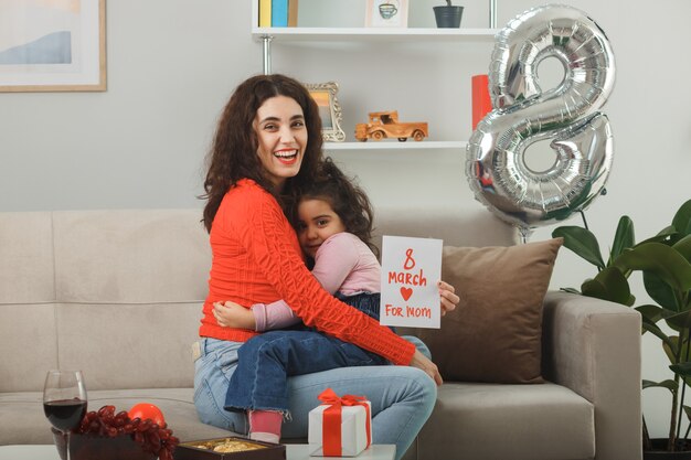 Madre felice con la sua piccola figlia seduta su un divano con in mano un biglietto di auguri sorridendo allegramente e abbracciando nel luminoso soggiorno che celebra la giornata internazionale della donna l'8 marzo
