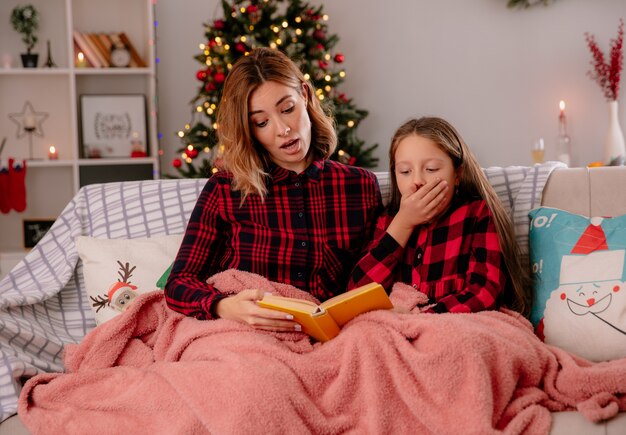 Madre scioccata e figlia che leggono il libro coperto di coperta che si siede sul divano e godersi il periodo natalizio a casa