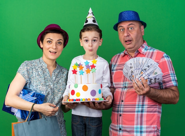 Madre sorpresa con cappello da festa viola che tiene in mano una scatola regalo e borse della spesa in piedi con il figlio che indossa un cappello da festa e tiene in mano una torta di compleanno con il padre che indossa un cappello da festa blu e tiene soldi