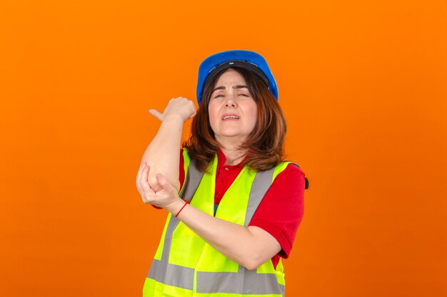 Maglia d'uso della costruzione dell'ingegnere della donna e casco di sicurezza che sembrano gomito commovente indisposto che ha dolore che controlla parete arancio isolata