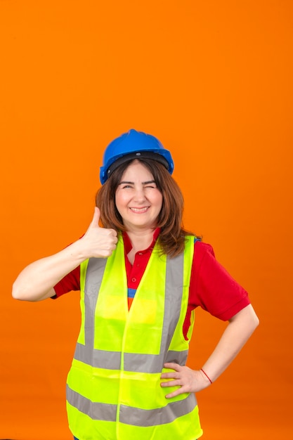 Maglia d'uso della costruzione dell'ingegnere della donna e sbattere le palpebre del casco di sicurezza che sorridono allegramente mostrando pollice su che controlla parete arancio isolata