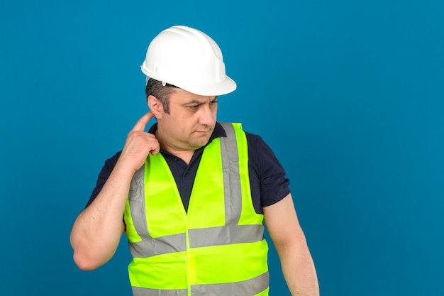 Maglia d'uso della costruzione gialla e casco di sicurezza d'uso dell'uomo invecchiato centrale che stanno con la testa commovente di sembrare pensieroso e che pensa sopra la parete blu isolata