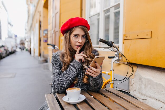 Magnifica giovane donna caucasica in abbigliamento elegante seduto con il telefono in street cafe