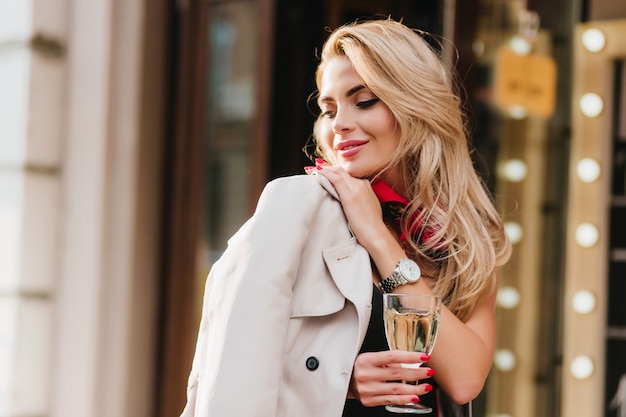 Magnifica giovane donna con acconciatura elegante che guarda lontano e sorridente in piedi. Ritratto all'aperto di ispirata signora dai capelli biondi con il manicure rosso che tiene il bicchiere di vino.