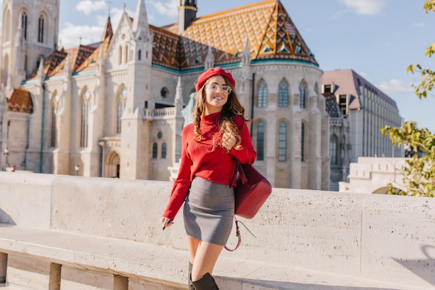 Magnifica ragazza magra in abbigliamento alla moda in piedi davanti al bellissimo palazzo nella soleggiata giornata di settembre