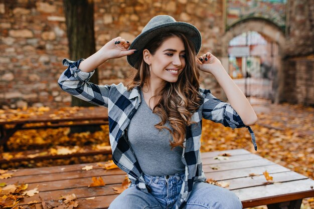 Magnifica ragazza pallida in cappello alla moda seduto sul tavolo nel parco e sorridente