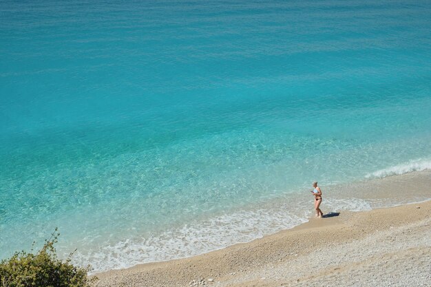 Magnifica vista della spiaggia sul Mar Egeo una bella donna adulta sta camminando lungo la spiaggia di pietra una luminosa giornata di sole Vacanza tropicale estiva sulla vista panoramica del Mar Egeo