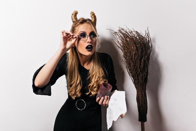 Mago femminile attraente che scherza durante il servizio fotografico di Halloween. Signora bionda caucasica in costume da strega in posa sul muro bianco.