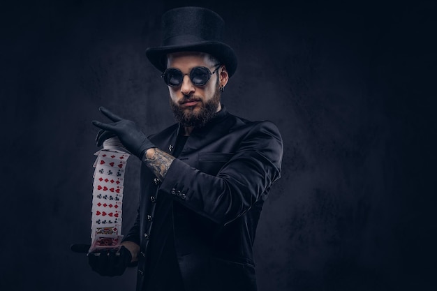 Mago in abito nero, occhiali da sole e cappello a cilindro, che mostra il trucco con le carte da gioco su uno sfondo scuro.
