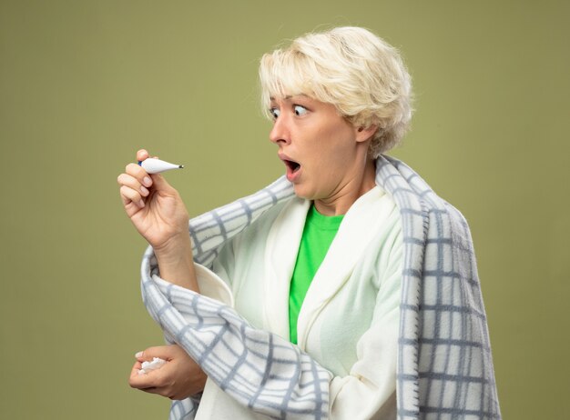 Malata malsana donna con i capelli corti avvolti in una coperta sensazione di malessere guardando il termometro essendo scioccato in piedi sopra la parete chiara