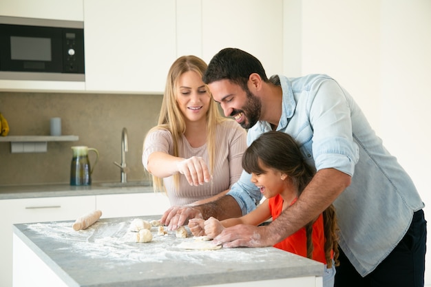 Mamma allegra e papà che insegnano alla figlia a fare la pasta sul tavolo della cucina con farina disordinata. Giovani coppie e la loro ragazza che cuociono insieme i panini o le torte. Concetto di cucina familiare