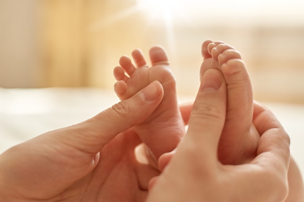 Mamma che fa massaggio al bambino, madre che massaggia il piede nudo del neonato, massaggio preventivo per il neonato, mamma che accarezza i piedi del bambino con entrambe le mani su sfondo chiaro.