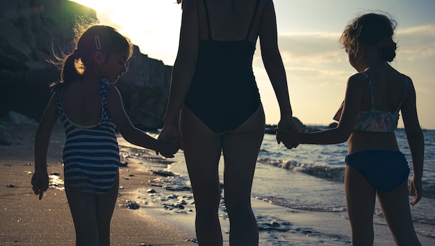 Mamma e due figlie piccole camminano lungo la riva del mare in costume da bagno, tenendosi per mano. Vee dal retro.