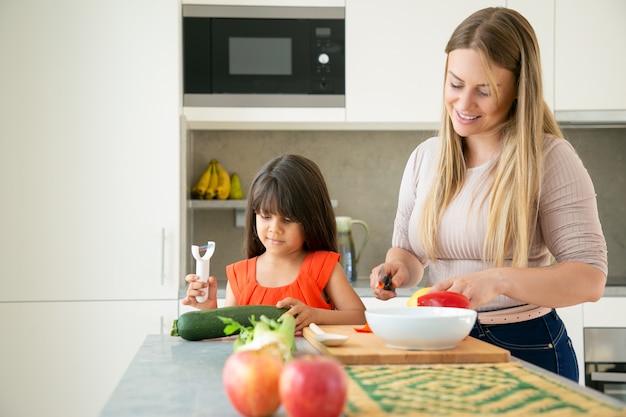 Mamma felice e figlia che cucinano insieme la cena. Ragazza e sua madre sbucciare e tagliare le verdure per insalata sul bancone della cucina. Concetto di cucina familiare