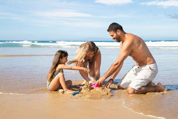 Mamma gioiosa, papà e figlia piccola che si godono le vacanze al mare insieme, giocano con i giocattoli della sabbia delle figlie, costruiscono castelli di sabbia