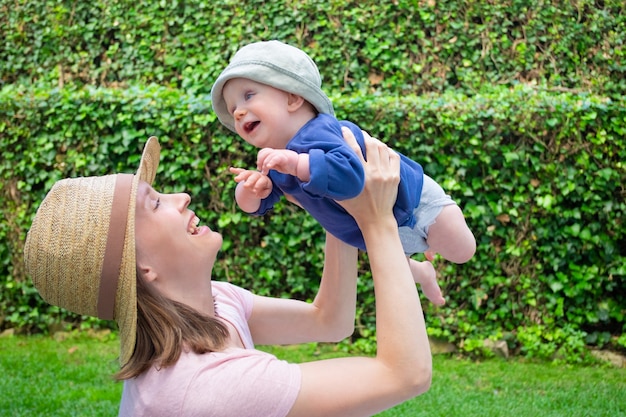 Mamma graziosa che gioca con la figlia nel parco e sorridente. Neonata sveglia in camicia blu e cappello che distoglie lo sguardo con la bocca aperta