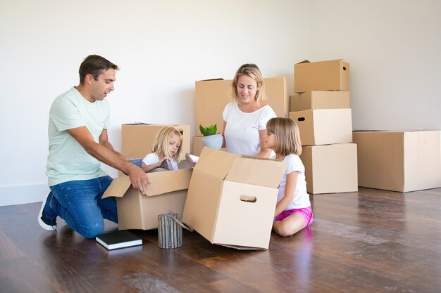 Mamma, papà e figlie piccole che disfano le cose nel nuovo appartamento, si siedono sul pavimento e prendono oggetti da scatole aperte