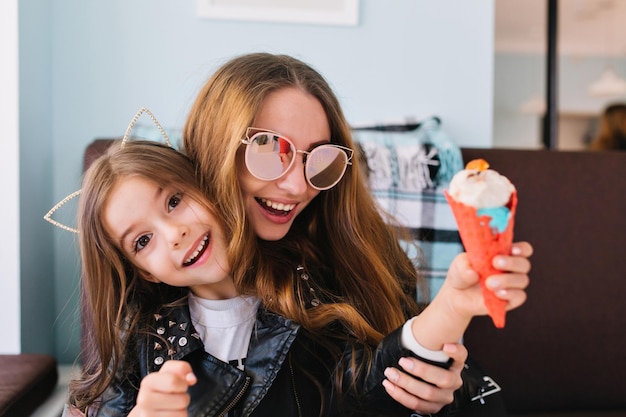 Mamma ridente alla moda in occhiali da sole che si diverte con la figlia eccitata al chiuso. Ritratto di una graziosa ragazza bruna che mangia un gustoso gelato seduto sulle ginocchia della madre mentre lei l'abbraccia dolcemente