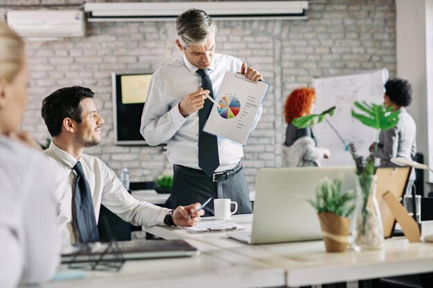 Manager di metà adulto che parla con il suo team di statistiche aziendali mentre mostra loro i dati grafici sulla chat a torta durante una riunione in ufficio