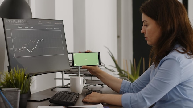 Manager donna che guarda lo schermo verde orizzontale sul telefono cellulare, lavorando in ufficio aziendale con display chroma key. Giovane donna che analizza lo schermo dello smartphone con mockup isolato.