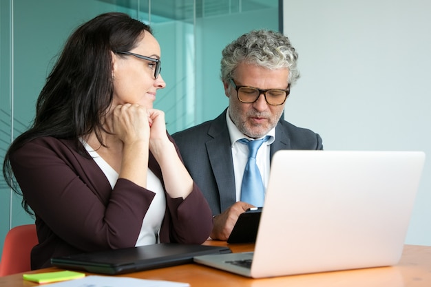 Manager e investitore che discutono del progetto di avvio. Colleghi riuniti a tavola con computer portatile aperto, utilizzando tablet e parlando.